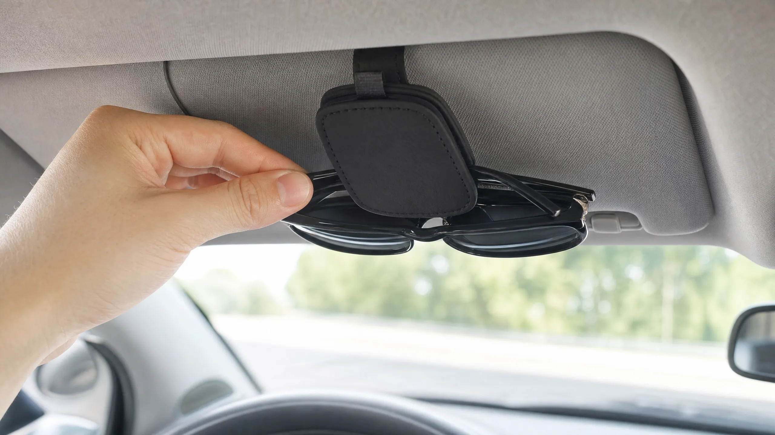 A person's hand placing or removing a pair of black sunglasses from a black magnetic leather holder attached to a car's sun visor.