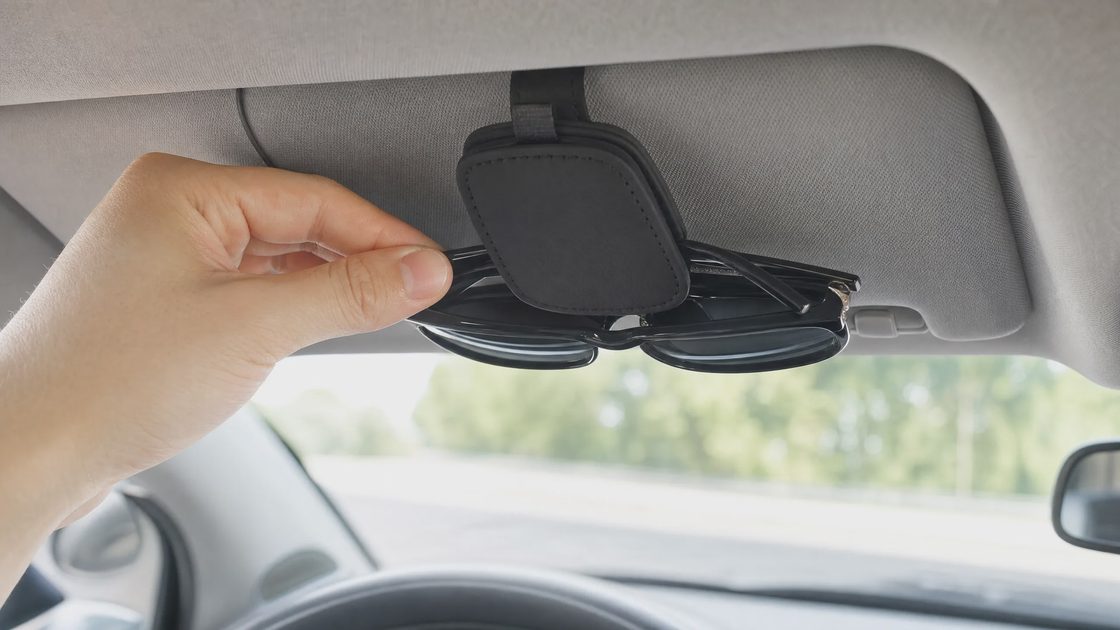 A person's hand placing or removing a pair of black sunglasses from a black magnetic leather holder attached to a car's sun visor.